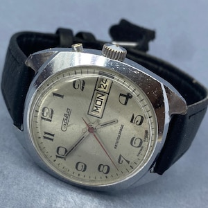Puede incluir: Un reloj de pulsera vintage plateado con una correa de cuero negro. La esfera del reloj muestra el día y la fecha, con "LUN 24" visible. El reloj tiene un bisel plateado y una esfera clara con números y agujas negras.