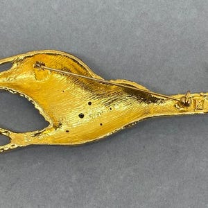 K&ouml;nnte beinhalten: Eine goldfarbene Brosche in Form eines stilisierten Vogels, m&ouml;glicherweise eines Roadrunners, mit ausgestreckten Beinen und einem langen Schnabel. Die Brosche hat einen Nadelverschluss auf der R&uuml;ckseite und besteht aus gl&auml;nzendem, strukturiertem Metall. Der Vogel liegt.