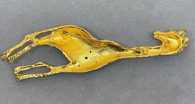 K&ouml;nnte beinhalten: Eine goldfarbene Brosche in Form eines stilisierten Vogels, m&ouml;glicherweise eines Roadrunners, mit ausgestreckten Beinen und einem langen Schnabel. Die Brosche hat einen Nadelverschluss auf der R&uuml;ckseite und besteht aus gl&auml;nzendem, strukturiertem Metall. Der Vogel liegt.
