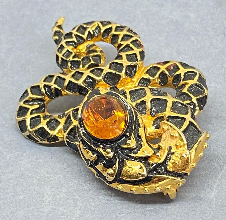 May include: A vintage snake brooch with a gold-tone body and black detailing. The snake's head features an orange gemstone. The brooch is a unique accessory, perfect for adding a touch of vintage flair to any outfit.