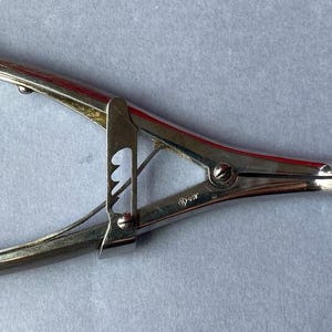 K&ouml;nnte beinhalten: Silberfarbenes chirurgisches Instrument mit gebogenen Griffen und gezahnter Spitze. Das Werkzeug hat ein Scharnierdesign mit Verriegelungsmechanismus und wird wahrscheinlich zum Greifen oder Klemmen verwendet.