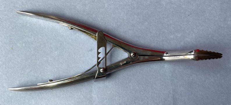 K&ouml;nnte beinhalten: Silberfarbenes chirurgisches Instrument mit gebogenen Griffen und gezahnter Spitze. Das Werkzeug hat ein Scharnierdesign mit Verriegelungsmechanismus und wird wahrscheinlich zum Greifen oder Klemmen verwendet.