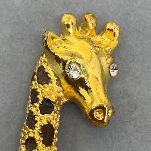 K&ouml;nnte beinhalten: Goldfarbene Giraffenbrosche mit detailliertem Design. Der Kopf der Giraffe hat zwei klare, funkelnde Strasssteine als Augen. Der Hals hat ein braunes und schwarzes Fleckenmuster.