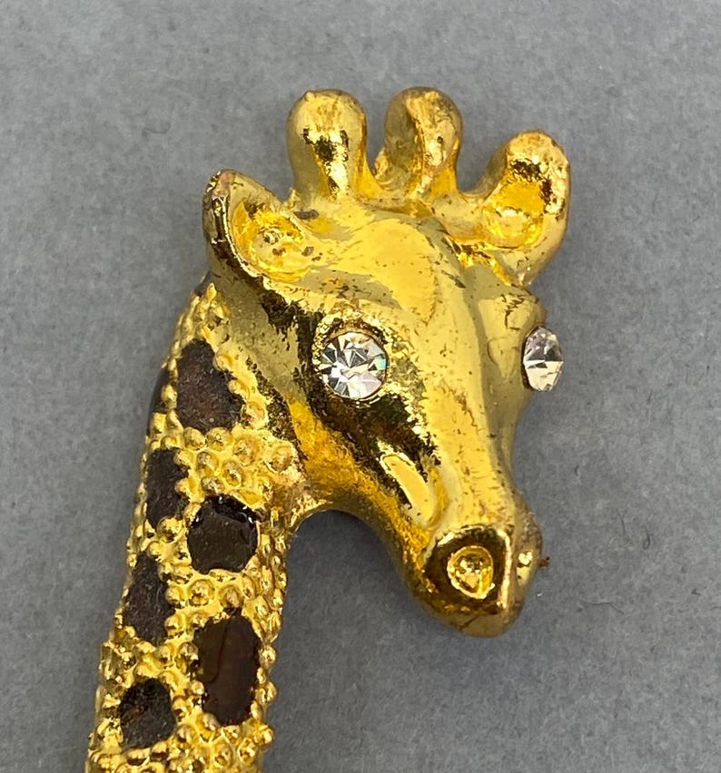 K&ouml;nnte beinhalten: Goldfarbene Giraffenbrosche mit detailliertem Design. Der Kopf der Giraffe hat zwei klare, funkelnde Strasssteine als Augen. Der Hals hat ein braunes und schwarzes Fleckenmuster.