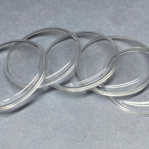 May include: A set of five clear, circular glass cabochons with a smooth, domed surface. Each piece is transparent and features a rounded edge, designed for crafting or jewelry making, presented as a group of individual circular glass components.