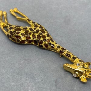 K&ouml;nnte beinhalten: Goldfarbene Giraffenbrosche mit braunen und goldenen Flecken. Der Kopf und die Beine der Giraffe sind mit klaren Kristallen verziert. Die Brosche ist ein dekoratives Accessoire, geeignet, um Kleidung oder Accessoires einen Hauch von Laune zu verleihen.