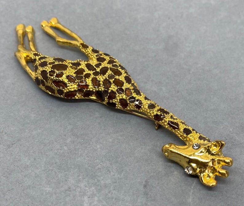 K&ouml;nnte beinhalten: Goldfarbene Giraffenbrosche mit braunen und goldenen Flecken. Der Kopf und die Beine der Giraffe sind mit klaren Kristallen verziert. Die Brosche ist ein dekoratives Accessoire, geeignet, um Kleidung oder Accessoires einen Hauch von Laune zu verleihen.