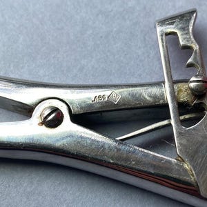 K&ouml;nnte beinhalten: Nahaufnahme eines Vintage-Werkzeugs aus silberfarbenem Metall mit Scharnierdesign. Das Werkzeug verf&uuml;gt &uuml;ber einen Hebel mit herzf&ouml;rmigem Ausschnitt und einer Reihe von Kerben. Eine kleine Markierung ist auf der Oberfl&auml;che des Werkzeugs sichtbar.