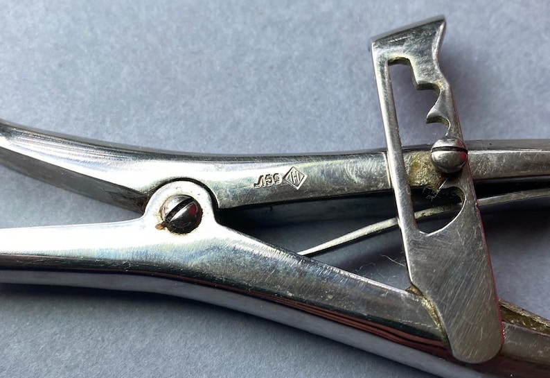 K&ouml;nnte beinhalten: Nahaufnahme eines Vintage-Werkzeugs aus silberfarbenem Metall mit Scharnierdesign. Das Werkzeug verf&uuml;gt &uuml;ber einen Hebel mit herzf&ouml;rmigem Ausschnitt und einer Reihe von Kerben. Eine kleine Markierung ist auf der Oberfl&auml;che des Werkzeugs sichtbar.