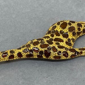 K&ouml;nnte beinhalten: Goldfarbene Giraffenbrosche mit braunen und dunkelroten Flecken. Die Giraffe liegt mit ausgestreckten Beinen. Ein kleiner, klarer Edelstein ist im Auge der Giraffe eingesetzt. Die Brosche ist ein dekoratives Accessoire.