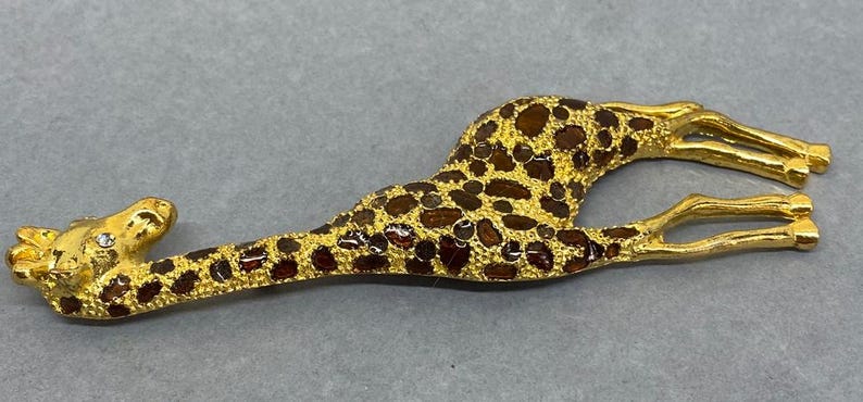 K&ouml;nnte beinhalten: Goldfarbene Giraffenbrosche mit braunen und dunkelroten Flecken. Die Giraffe liegt mit ausgestreckten Beinen. Ein kleiner, klarer Edelstein ist im Auge der Giraffe eingesetzt. Die Brosche ist ein dekoratives Accessoire.