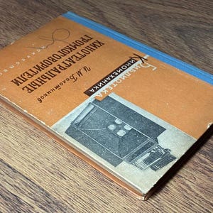 May include: A vintage book with a worn orange cover and blue spine. The cover features text in a foreign language and a black and white illustration of a machine. The book is titled "Kinoteatral'nye Gromkogovoriteli" and is on a wooden surface.