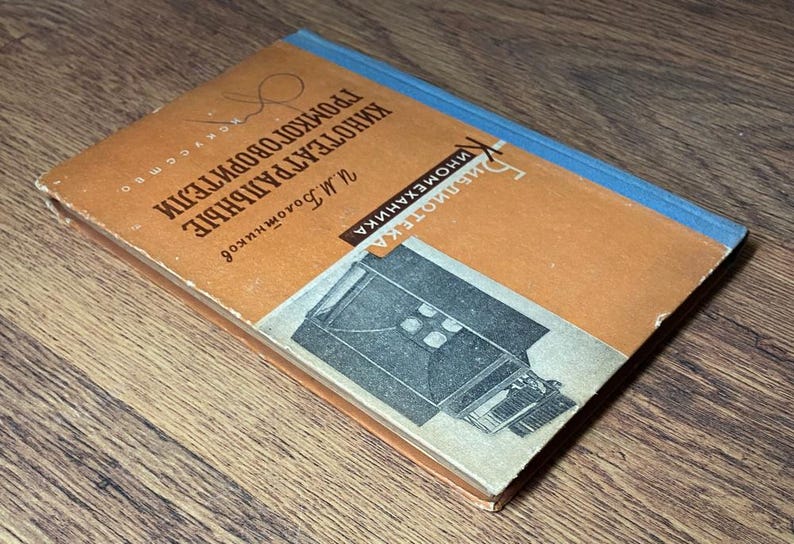 May include: A vintage book with a worn orange cover and blue spine. The cover features text in a foreign language and a black and white illustration of a machine. The book is titled "Kinoteatral'nye Gromkogovoriteli" and is on a wooden surface.