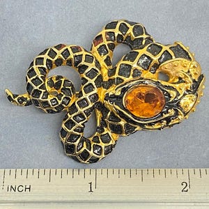 May include: A gold-toned snake brooch with black enamel detailing and an orange gemstone eye. The brooch is approximately 2 inches long, as measured by the inch ruler in the image. The snake's body is coiled, and the design is intricate.