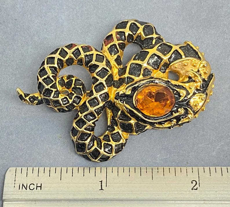 May include: A gold-toned snake brooch with black enamel detailing and an orange gemstone eye. The brooch is approximately 2 inches long, as measured by the inch ruler in the image. The snake's body is coiled, and the design is intricate.