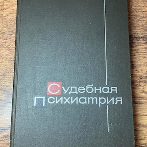 Può includere: Un libro grigio scuro con il titolo "Судебная психиатрия" in caratteri cirillici bianchi, con un logo quadrato rosso e bianco. Una linea bianca verticale corre lungo il lato destro. Il libro è su una superficie di legno.