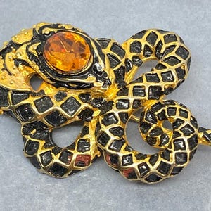 May include: A gold-toned snake brooch with black enamel detailing and a large, orange gemstone eye. The snake's body is intricately designed with a scale pattern, creating a striking and detailed accessory.