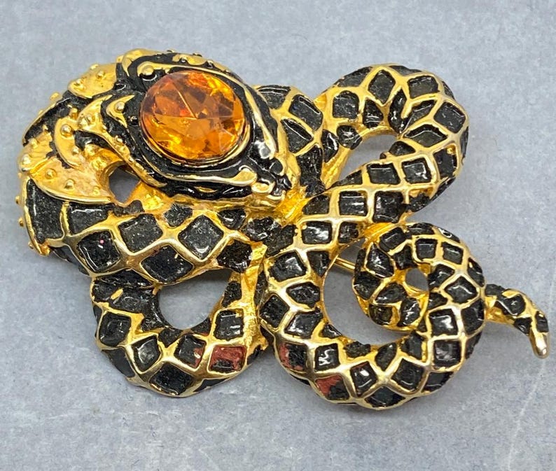May include: A gold-toned snake brooch with black enamel detailing and a large, orange gemstone eye. The snake's body is intricately designed with a scale pattern, creating a striking and detailed accessory.