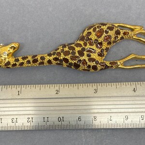 K&ouml;nnte beinhalten: Eine goldfarbene Giraffenbrosche mit braunen Flecken und einem Strassauge. Die Brosche ist etwa 10 cm lang, gemessen mit dem Lineal im Bild. Die Giraffe liegt mit angewinkelten Beinen.
