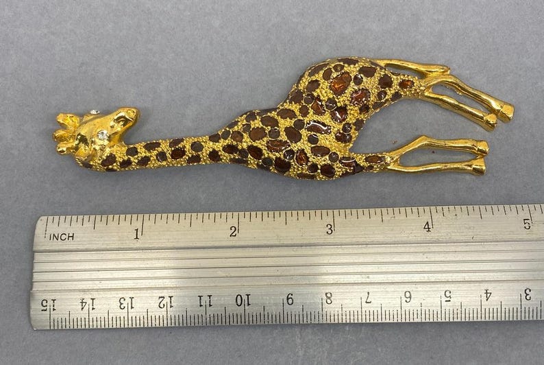 K&ouml;nnte beinhalten: Eine goldfarbene Giraffenbrosche mit braunen Flecken und einem Strassauge. Die Brosche ist etwa 10 cm lang, gemessen mit dem Lineal im Bild. Die Giraffe liegt mit angewinkelten Beinen.