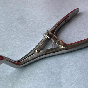 K&ouml;nnte beinhalten: Silberfarbenes medizinisches Instrument mit gebogenem Griff und zwei gegen&uuml;berliegenden Klingen. Das Instrument hat einen Scharniermechanismus und einen strukturierten Griff. Die Klingen haben eine rote Innenseite.