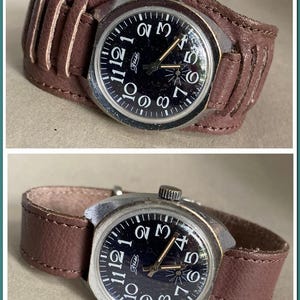 Puede incluir: Un reloj de pulsera vintage con esfera negra y números blancos, con una correa de cuero marrón estilo brazalete. La esfera del reloj tiene el texto "Jus" y agujas doradas. La correa es de cuero marrón.