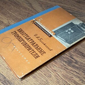 May include: Vintage book with an orange cover and blue spine. The cover features text in a foreign language, a graphic of a film camera, and a signature. The book is titled "Kino-teatral'nye Gromkogovoriteli".