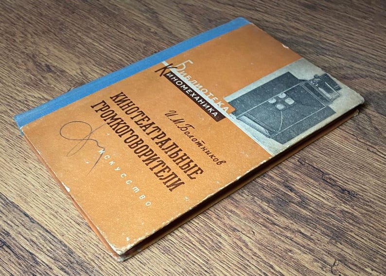 May include: Vintage book with an orange cover and blue spine. The cover features text in a foreign language, a graphic of a film camera, and a signature. The book is titled "Kino-teatral'nye Gromkogovoriteli".