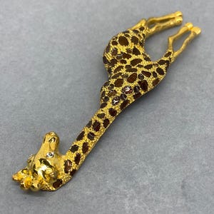 K&ouml;nnte beinhalten: Goldfarbene Giraffenbrosche mit braunen Flecken und funkelnden Akzenten. Der Kopf und die Beine der Giraffe sind detailliert, vor grauem Hintergrund. Dieses Schmuckst&uuml;ck mit Tiermotiv ist ein einzigartiges Accessoire.