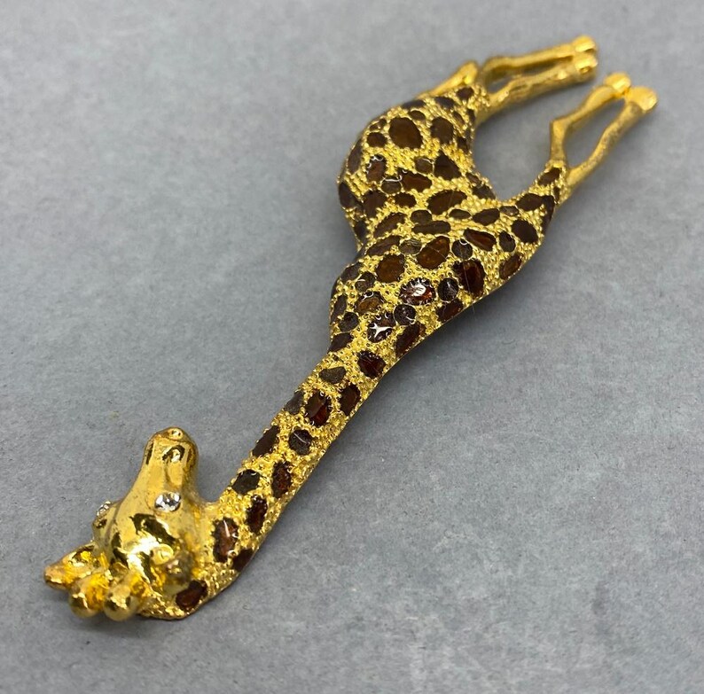 K&ouml;nnte beinhalten: Goldfarbene Giraffenbrosche mit braunen Flecken und funkelnden Akzenten. Der Kopf und die Beine der Giraffe sind detailliert, vor grauem Hintergrund. Dieses Schmuckst&uuml;ck mit Tiermotiv ist ein einzigartiges Accessoire.