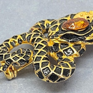 May include: Gold-toned snake brooch with black enamel detailing and a central amber-colored stone. The snake is coiled, with a detailed scale pattern. The brooch has a pin closure on the back, and is a vintage jewelry piece.