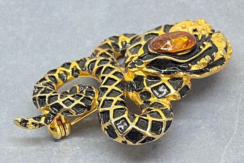 May include: Gold-toned snake brooch with black enamel detailing and a central amber-colored stone. The snake is coiled, with a detailed scale pattern. The brooch has a pin closure on the back, and is a vintage jewelry piece.