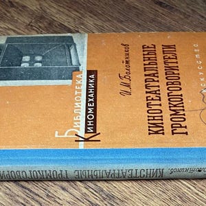 May include: Vintage book titled "Kinoteatralnye Gromkogovoriteli" in Russian. The cover features an illustration of a machine and text in orange and white. The spine is blue. The book is lying on a wooden surface.