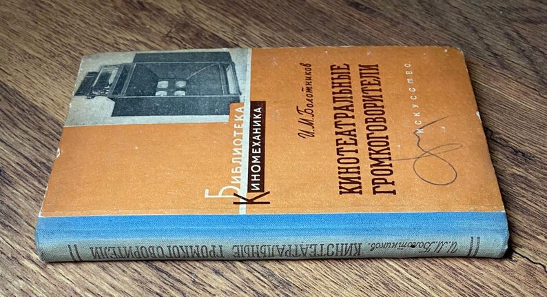 May include: Vintage book titled "Kinoteatralnye Gromkogovoriteli" in Russian. The cover features an illustration of a machine and text in orange and white. The spine is blue. The book is lying on a wooden surface.