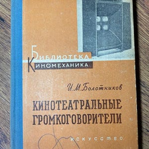 May include: Vintage book with an orange cover and blue spine. The cover features a black and white illustration of a speaker system and text in Russian. The book is titled "Kинотеатральные громкоговорители".