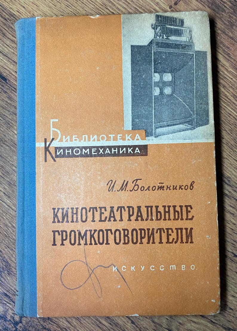 May include: Vintage book with an orange cover and blue spine. The cover features a black and white illustration of a speaker system and text in Russian. The book is titled "Kинотеатральные громкоговорители".
