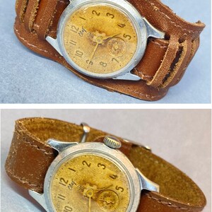 May include: Vintage wristwatch with a silver-toned case and a tan face, featuring black numerals and hands. The watch is attached to a wide, brown leather cuff-style band with decorative stitching.