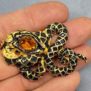 May include: A gold-toned snake brooch with black enamel detailing and a large, oval amber-colored stone. The snake's body is intricately designed with a scale pattern. The brooch is a vintage accessory.