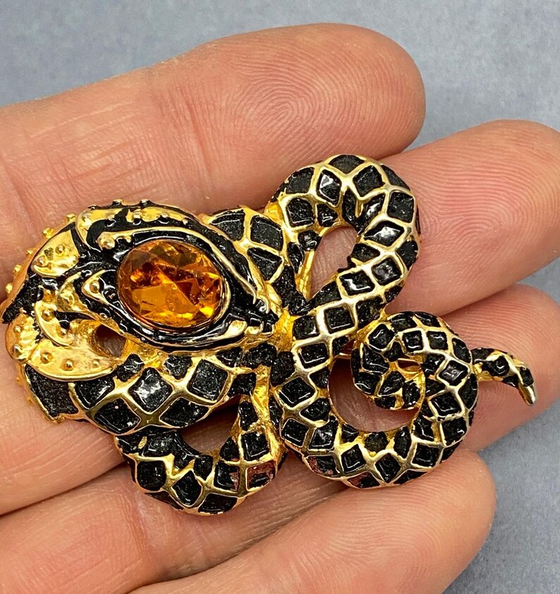 May include: A gold-toned snake brooch with black enamel detailing and a large, oval amber-colored stone. The snake's body is intricately designed with a scale pattern. The brooch is a vintage accessory.