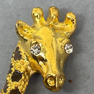 K&ouml;nnte beinhalten: Goldfarbene Giraffenbrosche mit Strassaugen und strukturiertem, geflecktem Hals. Der Kopf der Giraffe ist detailliert mit einer polierten Oberfl&auml;che, und die Brosche hat einen sicheren Nadelverschluss. Ein charmantes Accessoire mit Tiermotiv.