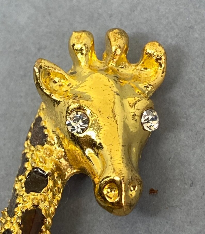 K&ouml;nnte beinhalten: Goldfarbene Giraffenbrosche mit Strassaugen und strukturiertem, geflecktem Hals. Der Kopf der Giraffe ist detailliert mit einer polierten Oberfl&auml;che, und die Brosche hat einen sicheren Nadelverschluss. Ein charmantes Accessoire mit Tiermotiv.