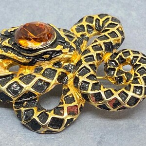 May include: A vintage snake brooch with a gold-tone metal base, black enamel detailing, and a large amber-colored gemstone. The snake is coiled, with a detailed scale pattern. The brooch is a unique piece of jewelry.