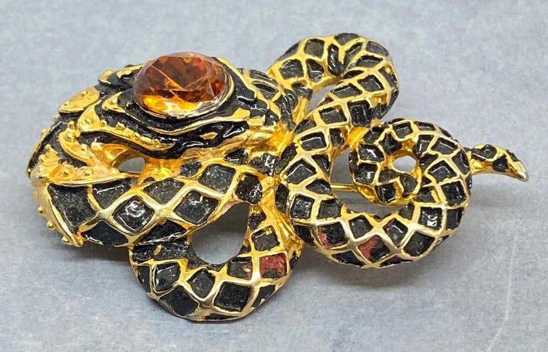 May include: A vintage snake brooch with a gold-tone metal base, black enamel detailing, and a large amber-colored gemstone. The snake is coiled, with a detailed scale pattern. The brooch is a unique piece of jewelry.