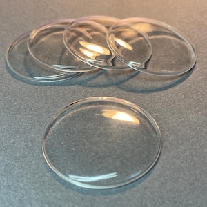 May include: A collection of five clear, circular, domed glass cabochons. Each piece features a smooth, transparent surface with a convex top and a flat bottom, designed for crafting or jewelry making. The items are arranged in a small cluster.