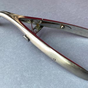 K&ouml;nnte beinhalten: Ein silberfarbenes chirurgisches Instrument mit einem gebogenen, geriffelten Ende und zwei langen, flachen Griffen. Das Instrument ist ge&ouml;ffnet und zeigt seinen inneren Mechanismus. Medizinisches Werkzeug.