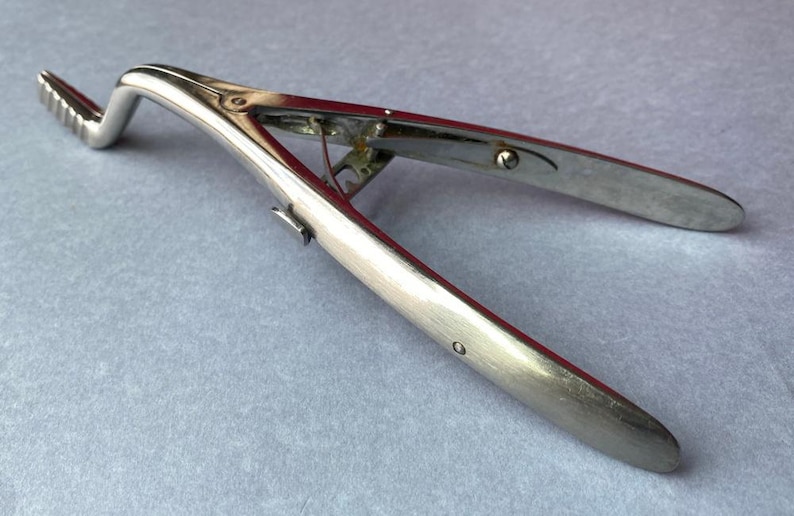 K&ouml;nnte beinhalten: Ein silberfarbenes chirurgisches Instrument mit einem gebogenen, geriffelten Ende und zwei langen, flachen Griffen. Das Instrument ist ge&ouml;ffnet und zeigt seinen inneren Mechanismus. Medizinisches Werkzeug.