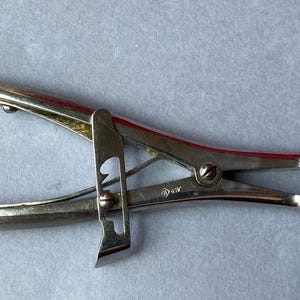 K&ouml;nnte beinhalten: Ein silberfarbenes chirurgisches Instrument mit Scharnierdesign. Das Werkzeug hat gebogene Griffe und gezahnte Spitzen. Das Instrument dient zum Klemmen oder Greifen von Objekten.