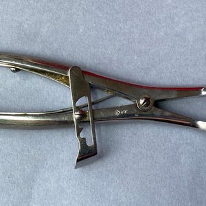 K&ouml;nnte beinhalten: Ein silberfarbenes chirurgisches Instrument mit gebogenen Backen und gezahnten Spitzen. Das Werkzeug hat ein Scharnierdesign mit einem Verriegelungsmechanismus und ist zum Greifen oder Halten von Objekten bestimmt. Das Instrument besteht aus Metall und wird wahrscheinlich in medizinischen oder wissenschaftlichen Bereichen verwendet.