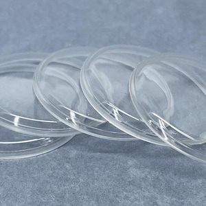 May include: A set of four clear, circular, slightly domed discs made of a smooth, transparent material. Each disc features a raised, rounded outer rim and a concave center, arranged in a slightly overlapping row on a flat surface.