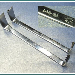 May include: Two silver-colored, curved metal tools, possibly surgical instruments, are displayed on a light gray surface. Each tool has a flat, rectangular end and a curved, hook-like end. A close-up shows markings including "B.M."
